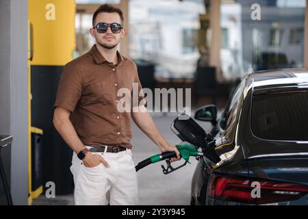 Lächelnder Mann, der sein Luxusauto an der Tankstelle betankt. Der Kerl guckt in die Kamera und gießt Benzin in den Tank eines modernen Fahrzeugs an der Tankstelle in der Stadt Stockfoto