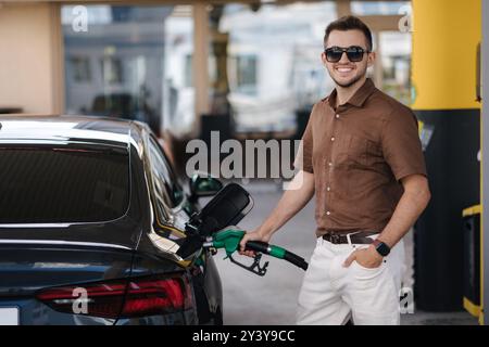 Lächelnder Mann, der sein Luxusauto an der Tankstelle betankt. Der Kerl guckt in die Kamera und gießt Benzin in den Tank eines modernen Fahrzeugs an der Tankstelle in der Stadt Stockfoto