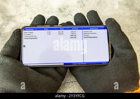 Ein Arbeiter, der Arbeitshandschuhe trägt und mit beiden Händen hält, einen digitalen Gehaltszettel auf einem Mobiltelefon, der eine Aufschlüsselung der Einnahmen anzeigt. Stockfoto