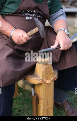 Rositz, Deutschland. September 2024. Ein Teilnehmer der 1. Mitteldeutschen Dengel-Meisterschaft arbeitet an seinem Werkstück. Bei Dengeling wird die Kante einer Sense mit Hammer und Amboss geschärft. Mehrere Dutzend Männer und Frauen aus Sachsen, Sachsen-Anhalt und Thüringen traten im Wettbewerb dieses alten Handwerks gegeneinander an, der im Rahmen der Rositzer Messe im Altenburger Land stattfand. Quelle: Sebastian Willnow/dpa/Alamy Live News Stockfoto