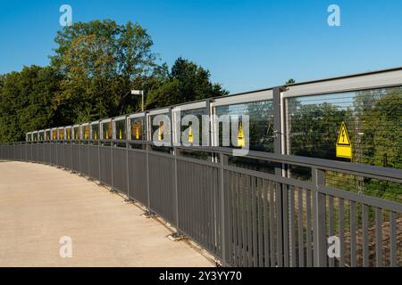 Ein gekrümmter Fußgängerweg weist mehrere Warnschilder entlang des Geländes auf, umgeben von Bäumen und blauem Himmel, was auf Sicherheitsaspekte in hinweist Stockfoto