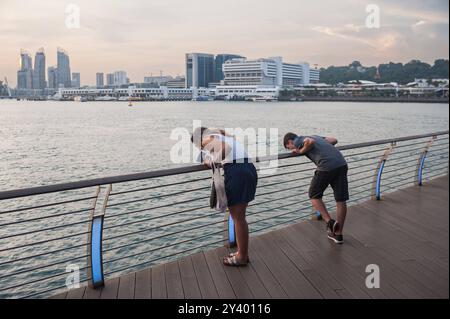 02.10.2016, Singapur, Republik Singapur, Asien - zwei Personen lehnen sich in der Abenddaemmerung auf der Uferpromenade des Sentosa Boardwalk uebers Gelaender, waehrend im Hintergrund Umrisse von Gebaeuden zu sehen sind. Der Boardwalk verbindet Hafenfront mit der Insel Sentosa. *** 02 10 2016, Singapur, Republik Singapur, Asien zwei Menschen lehnen sich in der Abenddämmerung über das Geländer an der Promenade Sentosa Boardwalk, während die Umrisse der Gebäude im Hintergrund zu sehen sind Stockfoto