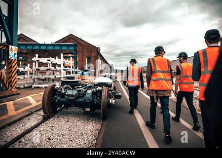 Gruppe von Arbeitnehmern, die in orangefarbenen Sicherheitswesten durch eine industrielle Eisenbahnanlage spazieren gehen und Stahlräder auf einem Zuggestell verfolgen Stockfoto