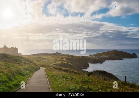 Ein malerischer Blick auf die Küste mit einem gewundenen Pfad, der zum Meer führt. Die Landschaft umfasst grasbewachsene Hügel, eine ferne Insel und einen bewölkten Himmel mit Stockfoto