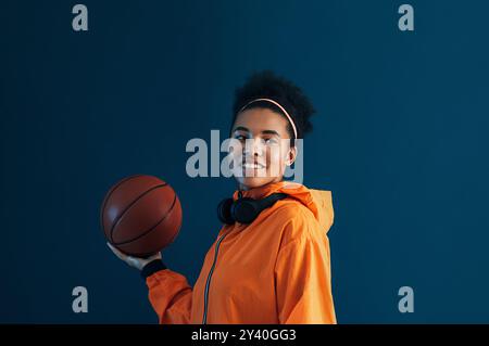 Junge lächelnde Frau, die Basketball an einer Hand hält und wegblickt, während sie über blauem Hintergrund steht. Positive Sportlerin in orangener Fitness-Kleidung Stockfoto