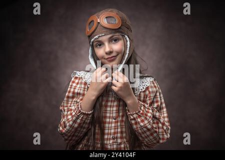 Retro-Porträt eines kaukasischen Mädchens in Vintage-Kleidung mit Fliegerhut vor einem alten braunen Tapetenhintergrund Stockfoto