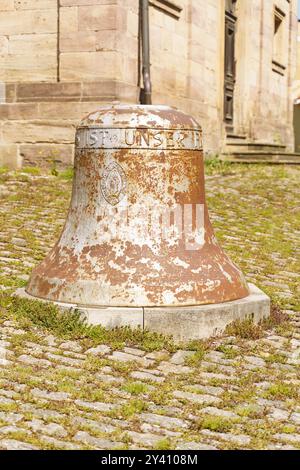 Waltershausen, Deutschland - 11. Juni 2023: Auf einem Kopfsteinpflasterweg steht Eine große, rostfarbene Glocke in einem charmanten europäischen Dorf, umgeben von Sonnenlicht Stockfoto