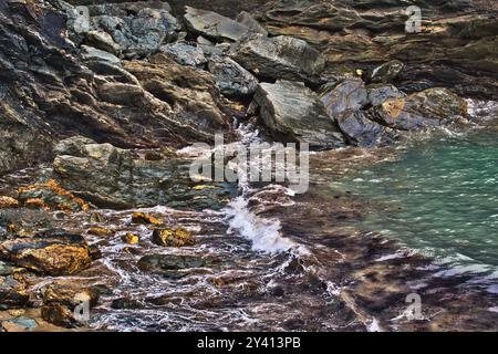 Eine felsige Küste mit Wellen, die sanft gegen die Steine prallen. Das Wasser ist klar und zeigt eine Mischung aus Farben von Felsen und Algen. Die Szene c Stockfoto