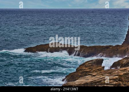 Ein malerischer Blick auf die felsige Küste mit Wellen, die gegen die Felsen stürzen, unter einem bewölkten Himmel. Das Meer ist tiefblau, was einen dramatischen Kontrast zum schafft Stockfoto