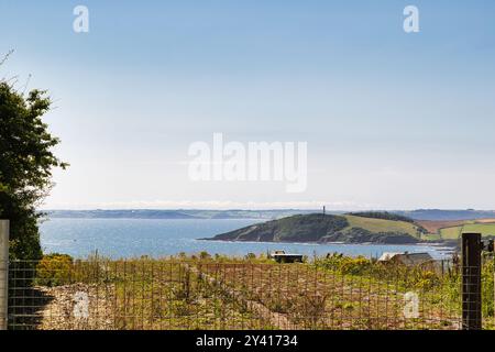 Ein malerischer Blick auf die Küste mit einem ruhigen Meer, fernen Hügeln und einem Leuchtturm auf einer kleinen Insel. Der Vordergrund zeigt grasbewachsenes Land mit karger Vegetation Stockfoto