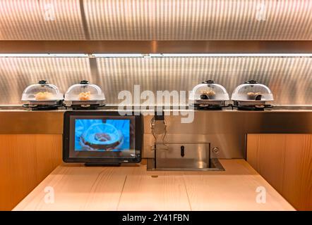 Vorderansicht eines Holztisches in einem japanischen Kaiten-zushi drehbaren Sushi-Bar-Restaurant mit einem Tablett als Menü und einem Förderband für Sushi p Stockfoto