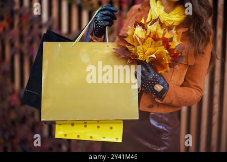 Hallo november. Nahaufnahme einer stilvollen Frau mittleren Alters in braunem Trenchcoat mit Einkaufstaschen und herbstgelben Blättern in der Stadt. Stockfoto
