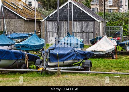 Eine Ansammlung von Booten, die mit blauen Planen bedeckt sind und auf einem grasbewachsenen Gelände in der Nähe eines Holzschuppen geparkt sind. Die Szene fängt eine ruhige maritime Atmosphäre mit einem Hi ein Stockfoto