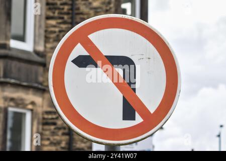 Ein kreisförmiges Verkehrsschild mit einem roten Rand und einem schwarzen Pfeil nach links, das anzeigt, dass keine Linkskurve zulässig ist. Das Schild ist an einem Pfosten in der Nähe eines Gebäudes angebracht Stockfoto