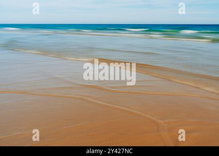 Sanfte Wellen erzeugen sanfte Muster an einem Sandstrand mit verschwommenem Bewegungseffekt. Stockfoto