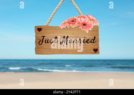 Flitterwochen. Holzbrett mit Worten, die gerade geheiratet wurden, und Blumen am Sandstrand Stockfoto