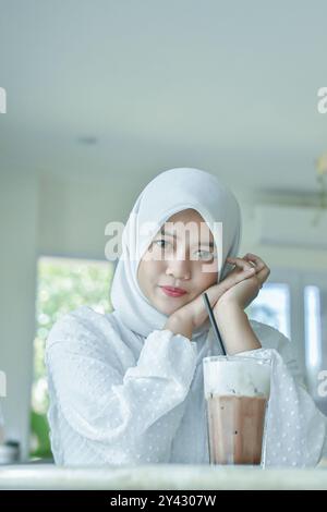 Pose einer jungen Frau, die einen weißen Hijab in einem Café trägt. Begleitet von einem Glas Eiskaffee Stockfoto