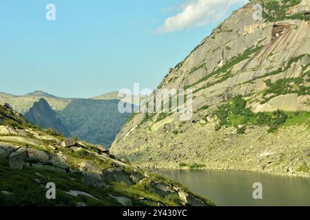 Ein hoher steiler Felsen am Ufer eines wunderschönen Bergsees mit felsigen Ufern, die an einem sonnigen Sommertag mit Gras und Moos bewachsen sind. Lake of Mountain Sp Stockfoto