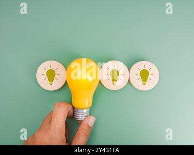 Leuchtendes Licht aus echter gelber Glühbirne, die von Hand gehalten wird, und Leuchtensymbole in rundem Holzblock, isoliert auf grünem Hintergrund, minimalistischer Stil. Mögliche Idee, Stockfoto