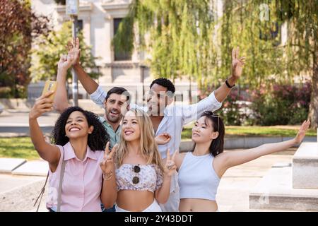 Fünf fröhliche Freunde treffen sich zu einem Selfie in einem sonnigen Park, lächeln, posieren mit Friedenszeichen und genießen einen lebhaften gemeinsamen Moment inmitten von Grün. Stockfoto