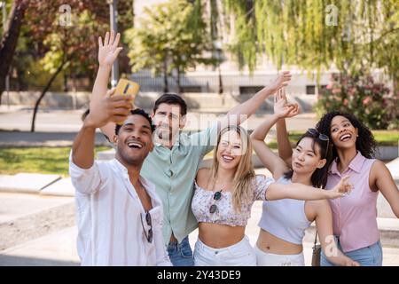 Eine lebhafte Gruppe von fünf Freunden macht an einem sonnigen Tag ein fröhliches Selfie im Freien, lächelt, winkt und posiert spielerisch in einem Park mit viel Grün. Stockfoto