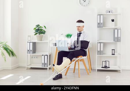 Behinderter Geschäftsmann, der im Büro mit einem Laptop arbeitet und auf einem Stuhl sitzt, mit einem bandagierten Kopf Stockfoto