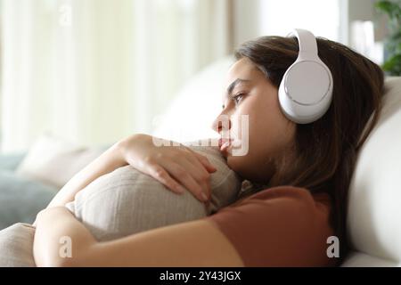 Traurige Frau mit Kopfhörern, die Musik hört und das Kissen umarmt, sitzt zu Hause auf einem Sofa Stockfoto