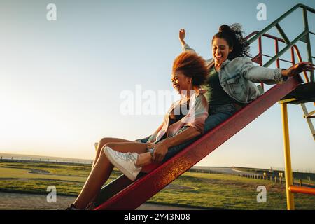 Zwei Freunde teilen sich einen unbeschwerten Moment auf einer Spielplatzrutsche während einer goldenen Stunde Sonnenuntergang. Das Bild fängt die Essenz von Spaß, Freiheit und Gesellschaft ein Stockfoto