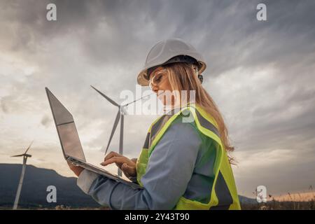 Eine erfahrene Ingenieurin führt wichtige Forschungen in einem modernen Windpark durch und nutzt fortschrittliche Technologien auf ihrem Laptop, um die Nachhaltigkeit zu verbessern und zu verbessern Stockfoto