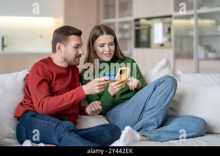 Online-Shopping zu Hause. Interessierte Paare bezahlen für Einkäufe mit Bankkarte, das Telefon sitzt auf der Couch. Stockfoto