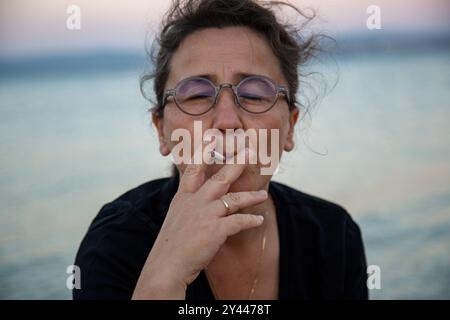 Frau mittleren Alters mit Brille raucht am Strand bei Sonnenuntergang Stockfoto