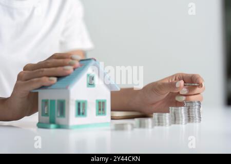 Person zählt Münzen neben einem kleinen Modellhaus auf einem weißen Tisch, Konzept, Geld für Immobilieninvestitionen zu sparen Stockfoto