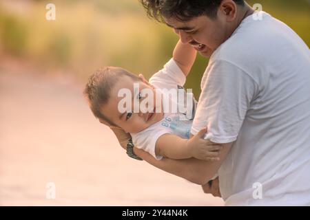 Vater, der den Jungen wiegt und der Junge, der vor der Kamera lächelt. Vater und kleiner Junge tragen weiße T-Shirts. Foto mit natürlichem Hintergrund und Sonnenlicht im Th Stockfoto