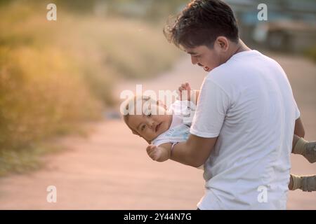 Vater hält und schwingt Baby Junge und Baby Junge mit unschuldigen Ausdruck der Überraschung. Vater und kleiner Junge tragen weiße T-Shirts. Stockfoto