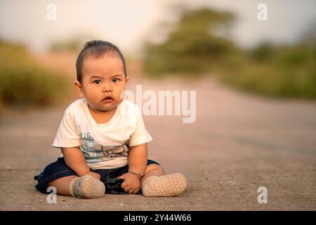 Süßes asiatisches Baby, das auf der Straße sitzt. Junge, der vor die Kamera blickt und ein weißes Hemd trägt. Fotoatmosphäre am Nachmittag. Stockfoto