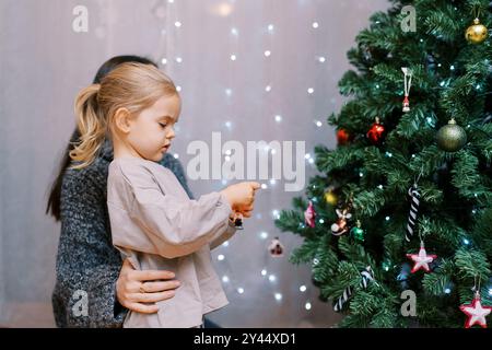 Mom hält ein kleines Mädchen an der Taille, während sie Spielzeug an den Weihnachtsbaum hängt. Seitenansicht Stockfoto