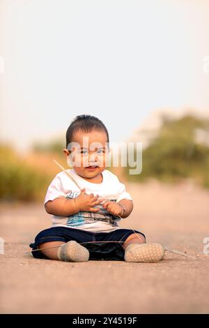 Süßes asiatisches Baby, das auf der Straße sitzt. Junge, der wildes Gras hält, während er süß lächelt und ein weißes Hemd in Richtung Kamera trägt. Fotoatmosphäre in t Stockfoto