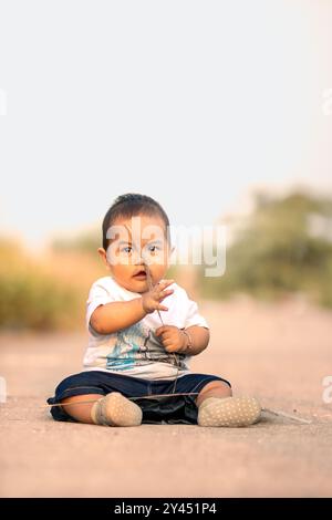 Süßes asiatisches Baby, das auf der Straße sitzt. Junge, der wildes Gras hält, während er süß lächelt und ein weißes Hemd in Richtung Kamera trägt. Fotoatmosphäre in t Stockfoto