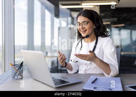 Eine junge Ärztin mit einem Headset, die mit einem Patienten spricht, ein Klinikmitarbeiter berät aus der Ferne, Online-Support, eine Inderin benutzt einen Laptop, lächelt glücklich und spricht über die Krankheit des Kunden. Stockfoto