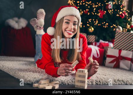 Foto in voller Länge von fröhlichem Mädchen liegend Boden Spiel Jenga gekleidet rot weihnachtskleidung Tradition Atmosphäre Dekor Vibe Haus Indoor Zimmer Stockfoto