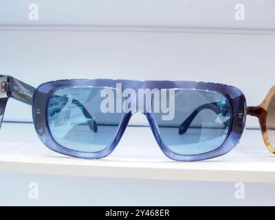 Blaue Sonnenbrillen mit dicken Rahmen und blauen Gläsern sitzen auf einem weißen Regal und spiegeln das Innere des Büros eines Optikers wider Stockfoto