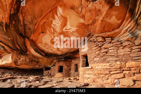 Gefallene Dachruinen, eine alte Steinkammer, die unter einer massiven überhängenden Felsformation mit komplexen Rot- und Orangetönen errichtet wurde. Cedar Mesa, Utah Stockfoto