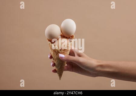 Hand hält Waffelkegel mit mehreren Hühnereiern auf beigem Hintergrund, Hühnerfarm Konzept, Eiermarkt Stockfoto