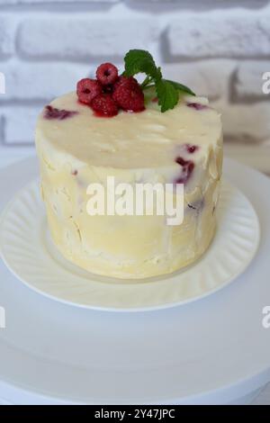 Hausgemachter Kuchen bedeckt mit weißer Schokolade und dekoriert mit roten FrühlingsHimbeeren, grünem Minzblatt unperfekt backen, Gebäck zu Hause bunter saftiger Kuchen und frischen Beeren auf weißer Platte Stockfoto