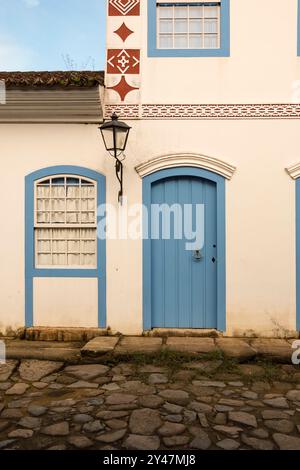 Paraty, Brasilien. Fassade eines Kolonialhauses aus dem Jahr 1836 in weiß-blauen Farben. Tür, Fenster und alte Straßenlaterne. Hintergrund blauer Himmel. Stockfoto