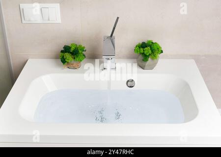 Wasser strömt aus einem Chromhahn in ein weißes Waschbecken aus Nahaufnahme, das Konzept, Wasserressourcen zu sparen und natürliche Ressourcen zu schützen. Stockfoto