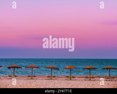 Eine ruhige Strandszene mit mehreren Strohschirmen, die an einem mit Kieselsteinen übersäten Ufer vor dem Sonnenuntergangshimmel aufgereiht sind, Altea, Spanien Stockfoto