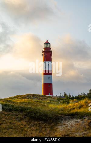 Der Leuchtturm ragt über sanften Sanddünen, wenn die Sonne untergeht, und wirft ein warmes Leuchten auf die Landschaft. Die roten und weißen Streifen sorgen für einen lebendigen Kontrast Stockfoto