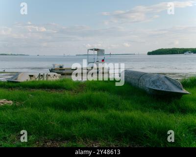 Weitwinkelblick über das Metallpontonrohr, das an Land in grünem Gras gespült wird, zum Krabbenanglerboot mit roten Schwimmbojen. Über Strand und Ebbe Stockfoto