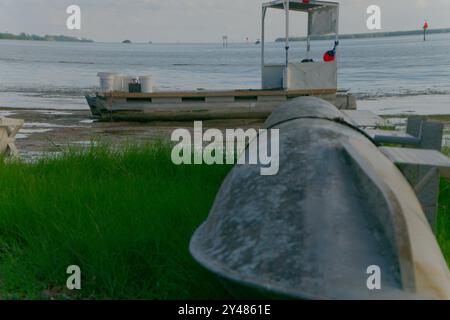 Weitwinkelblick über das Metallpontonrohr, das an Land in grünem Gras gespült wird, zum Krabbenanglerboot mit roten Schwimmbojen. Über Strand und Ebbe Stockfoto